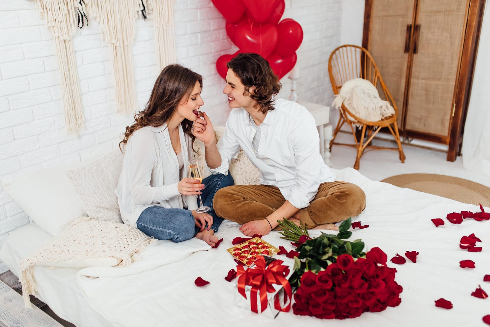 handsome man feeding girlfriend with chocolate candy drinking champagne while sitting bed home st valentines day for busy parents