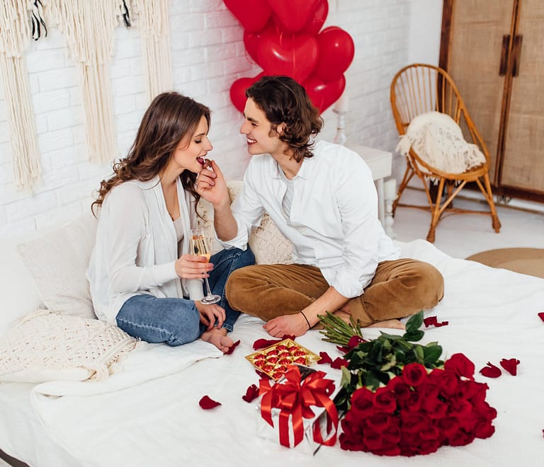 Handsome man feeding girlfriend with chocolate candy drinking champagne while sitting bed home on Valentine's Day