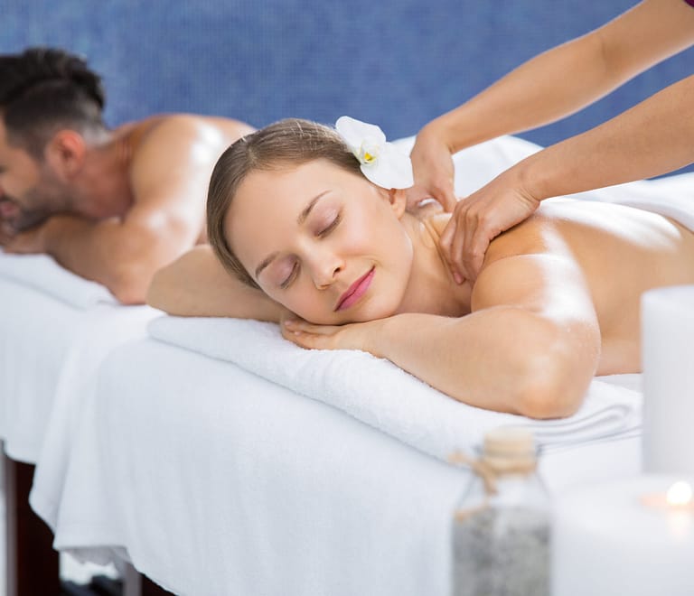 Couple getting massage together in spa and wellness center. Young Caucasian woman with flower in hair getting back massage. Man lying on massage table and relaxing after Valentine's day treatment in background