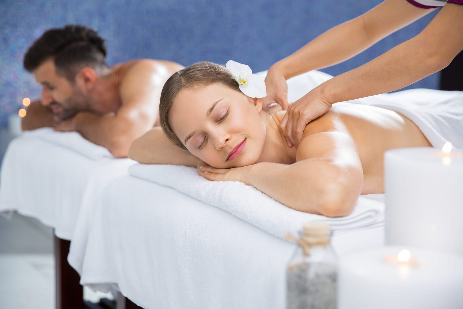 Couple getting massage together in spa and wellness center. Young Caucasian woman with flower in hair getting back massage. Man lying on massage table and relaxing after treatment in background